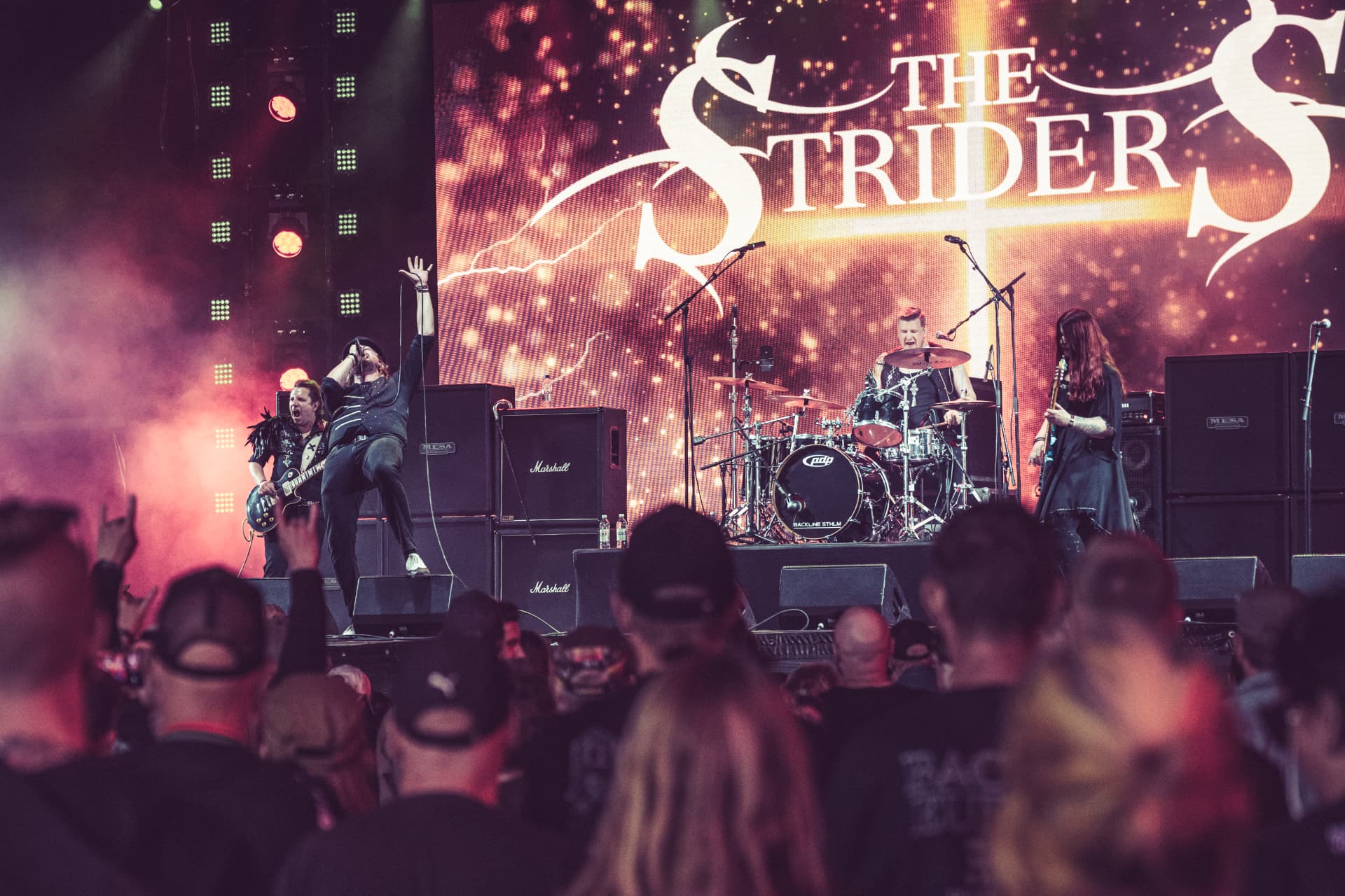 The Striders live on stage in front of a big screen with the band logo